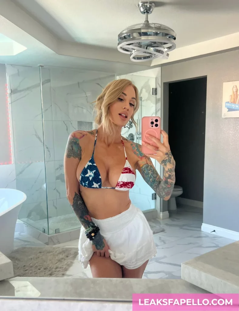 Sarah Jessie, Thick Blonde Tattooed Big tits OnlyFans model, is wearing a bikini with US flag design and a white cover up, mirror shot inside the bathroom