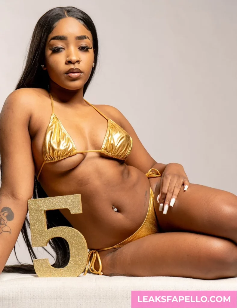 Angel Ace Pierce, ebony tattooed OnlyFans model, wearing a go;d colored lingerie, long sitting on the floor with a gold colored number 5 on the side