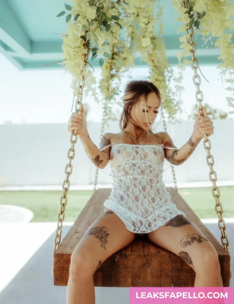 Linda Lan, Blonde Big Tits Curvy Tattooed OnlyFans model, is wearing a white dress, witting at the edge of the swing