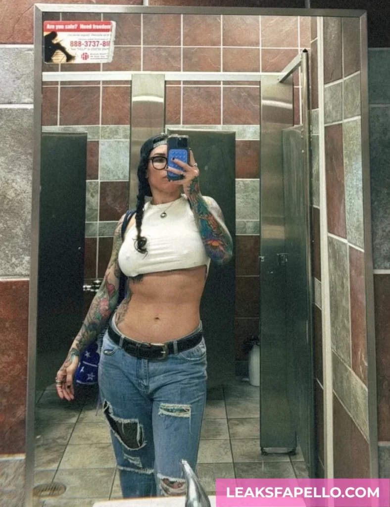 Bianca Bailee, thick curvy tattooed OnlyFans model, wearing a white crop top and ripped jeans, having a mirror selfie inside a public restroom